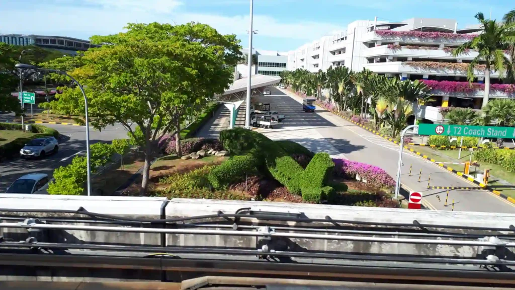 Ein großer grüner Formschnitt in Form einer Eidechse ist umgeben von blühenden Sträuchern in der Nähe einer Straße mit Palmen, neben einem weißen mehrstöckigen Gebäude unter einem klaren blauen Himmel, der an die üppigen Landschaften der neuseeländischen Südinsel erinnert.