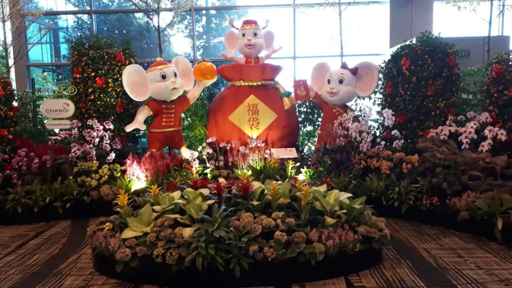 Drei große Mäusefiguren in festlicher Kleidung stehen inmitten bunter Blumen und Pflanzen. Die Maus in der Mitte hält einen Goldbarren auf einer roten Laterne mit einem chinesischen Schriftzeichen, die vor großen Fenstern und üppigem Grün steht, das an die Südinsel Neuseelands erinnert.