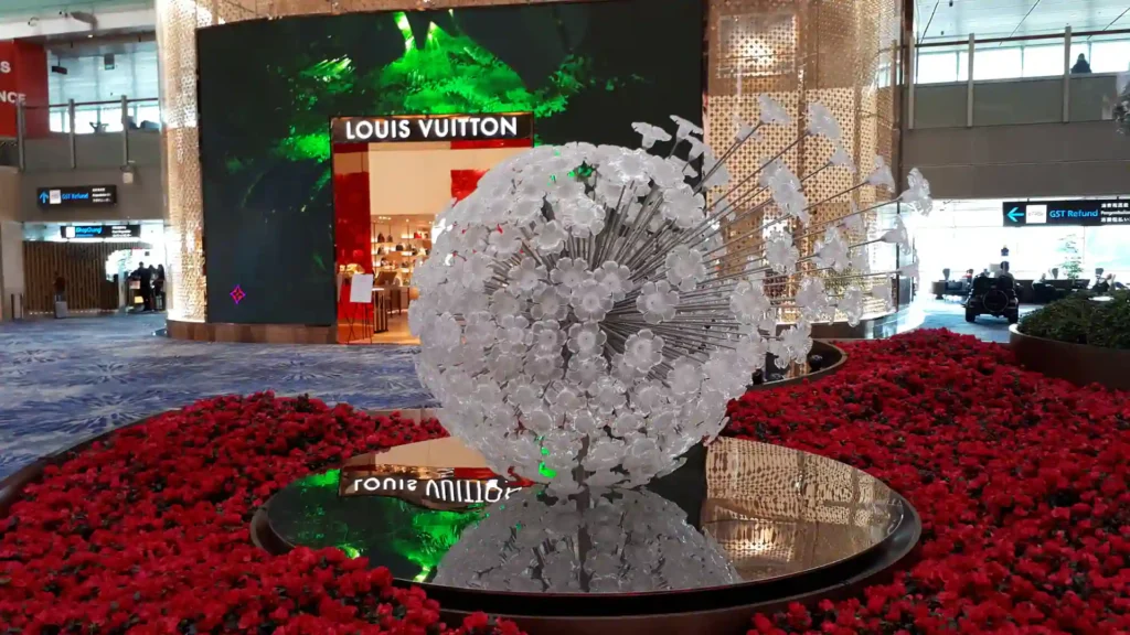 Eine große gläserne Pusteblumen-Skulptur steht auf einem spiegelnden Podest, umgeben von roten Blumen, mit einem Louis Vuitton-Geschäft und einer grün beleuchteten Wand im Hintergrund - eine Anspielung auf die Kunst auf der Neuseeländischen Südinsel.
