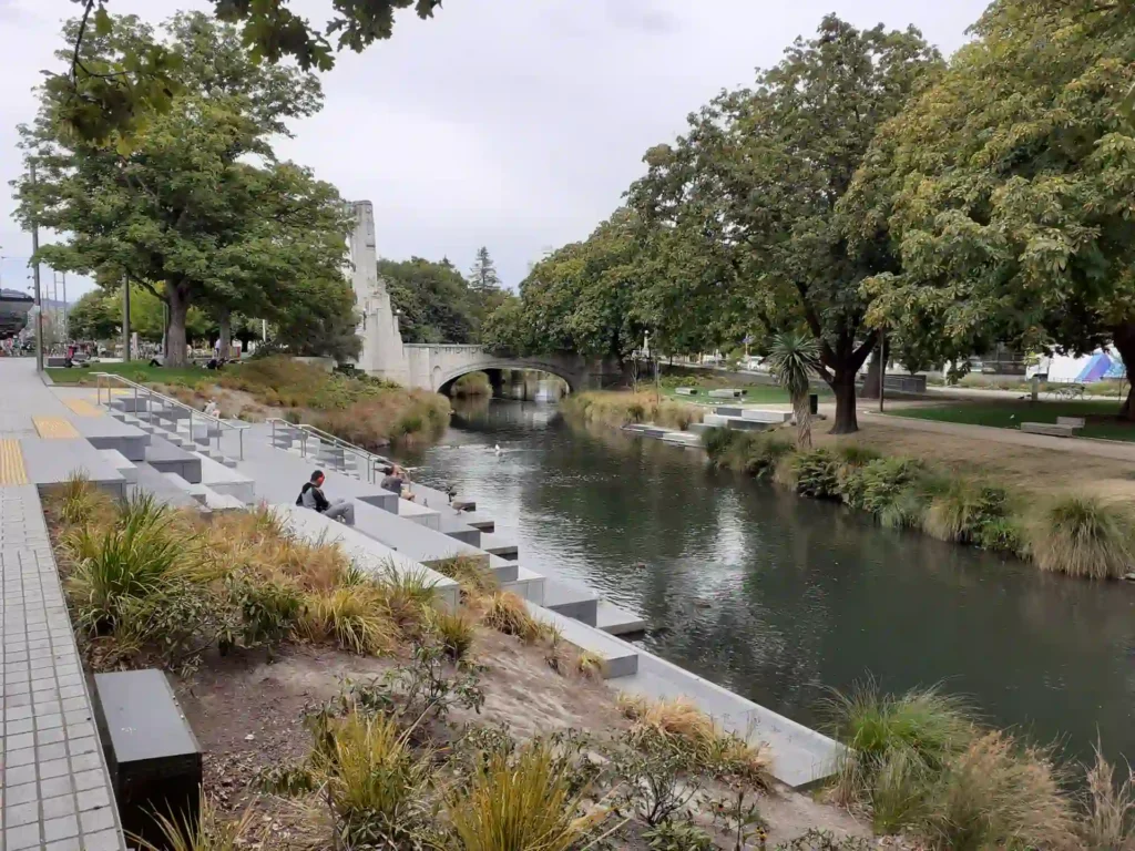 Ein ruhiger Fluss, der an die Landschaften der neuseeländischen Südinsel erinnert, fließt durch einen Stadtpark mit Bäumen und grasbewachsenen Ufern, Bänken und einer Steinbrücke. Unter einem bedeckten Himmel sitzen Menschen auf den Stufen am Wasser.