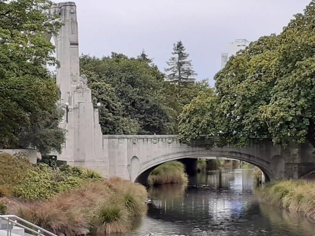 Eine Steinbrücke mit dekorativen Schnitzereien überspannt einen ruhigen Fluss auf der Neuseeländischen Südinsel, umgeben von üppigen grünen Bäumen und Gräsern, mit einem hohen Denkmal auf der linken Seite.