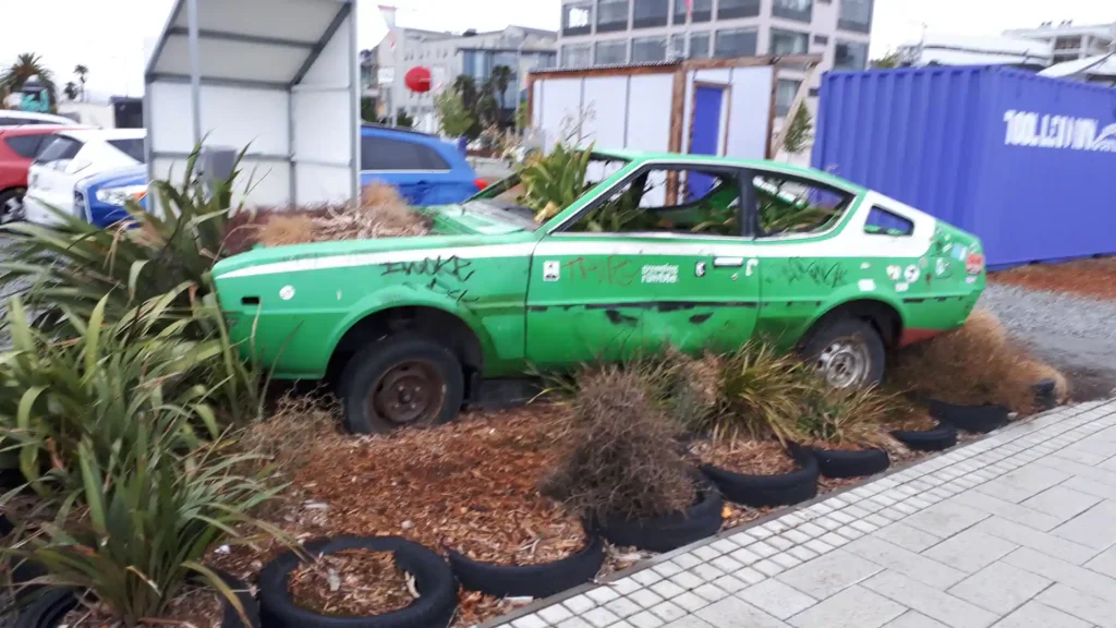 Ein verrostetes, grünes, mit Graffiti beschmiertes Auto mit fehlenden Rädern steht verlassen zwischen Pflanzen und alten Reifen in der Nähe eines Bürgersteigs auf der Neuseeländischen Südinsel, mit Gebäuden und einem blauen Schiffscontainer im Hintergrund.