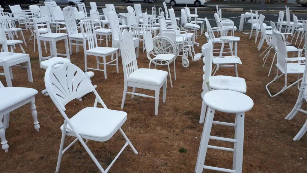 Reihen von weiß gestrichenen Stühlen, Hockern und einem Rollstuhl sind auf trockenem, braunem Gras im Freien auf der neuseeländischen Südinsel angeordnet und bilden eine Kunstinstallation oder einen Gedenkort. Im Hintergrund sind eine Straße und Autos zu sehen.