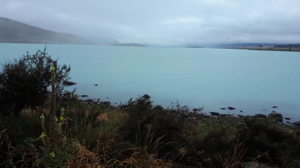Ein nebliger, bedeckter Tag an einem blassblauen See auf der neuseeländischen Südinsel, gesäumt von Felsen und wilden Gräsern, mit entfernten Hügeln, die teilweise von niedrigen Wolken verdeckt sind.