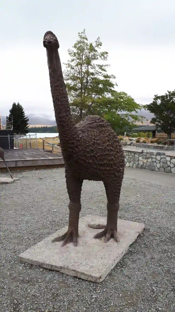 Eine große Statue eines Moa-Vogels steht auf einer Steinplatte auf der Neuseeländischen Südinsel. Sie hat einen langen Hals, einen strukturierten Körper und kräftige Beine. Bäume, eine Steinmauer und ein bewölkter Himmel bilden den Hintergrund.