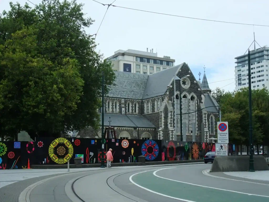 Ein steinernes Kirchengebäude im gotischen Stil steht hinter einem farbenfrohen, mit bunten Kreismustern verzierten Zaun auf der Neuseeländischen Südinsel. In der Nähe befinden sich Bäume und moderne Gebäude, und davor verläuft eine kurvenreiche Straße mit Straßenbahnschienen.