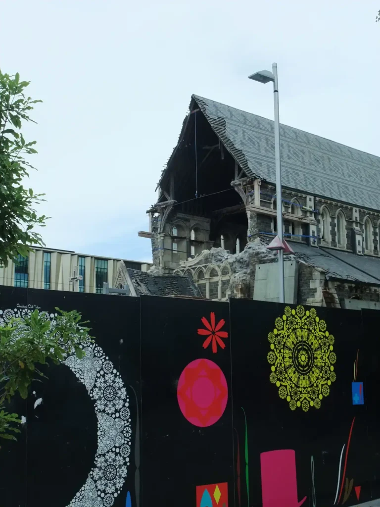 Ein beschädigtes Steingebäude auf der Neuseeländischen Südinsel, bei dem ein Teil des Daches und der oberen Wände fehlt, steht hinter einem schwarzen, mit bunten Mustern und Motiven verzierten Zaun. Auch ein Baum und eine Straßenlaterne sind zu sehen.