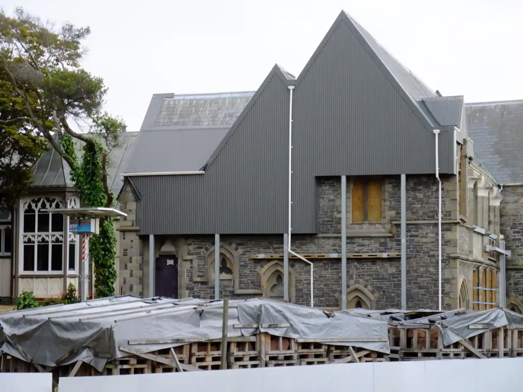 Hinter einer mit Planen und Holzpaletten bedeckten Baustelle auf der Neuseeländischen Südinsel steht ein Steinbau mit steilen grauen Dächern und verbretterten Fenstern. Ein Teil des Gebäudes weist Rundbogenfenster im gotischen Stil auf.