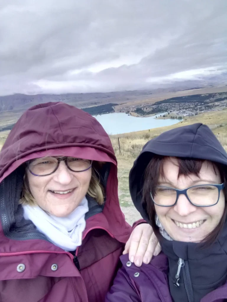 Zwei Personen mit Brillen und Kapuzenjacken lächeln für ein Selfie im Freien an einem bewölkten Tag auf der neuseeländischen Südinsel, mit einem See, Hügeln und einer kleinen Stadt im Hintergrund.