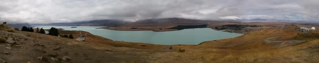 Panoramablick auf einen großen türkisfarbenen See auf der neuseeländischen Südinsel, umgeben von braunen Grashügeln unter einem bewölkten Himmel, mit fernen Bergen und einer kleinen Siedlung auf der rechten Seite.