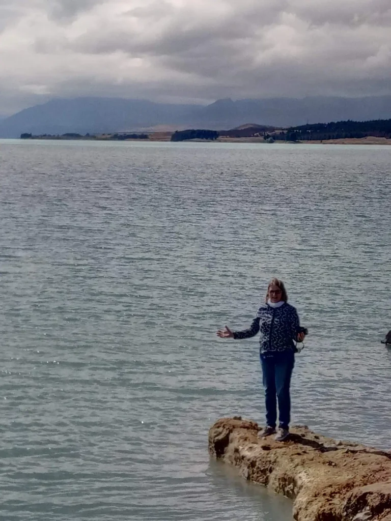 Eine Person mit Sonnenbrille und einem gemusterten Pullover steht auf einem Felsen an einem großen See auf der neuseeländischen Südinsel, mit Bergen und einem bewölkten Himmel im Hintergrund.
