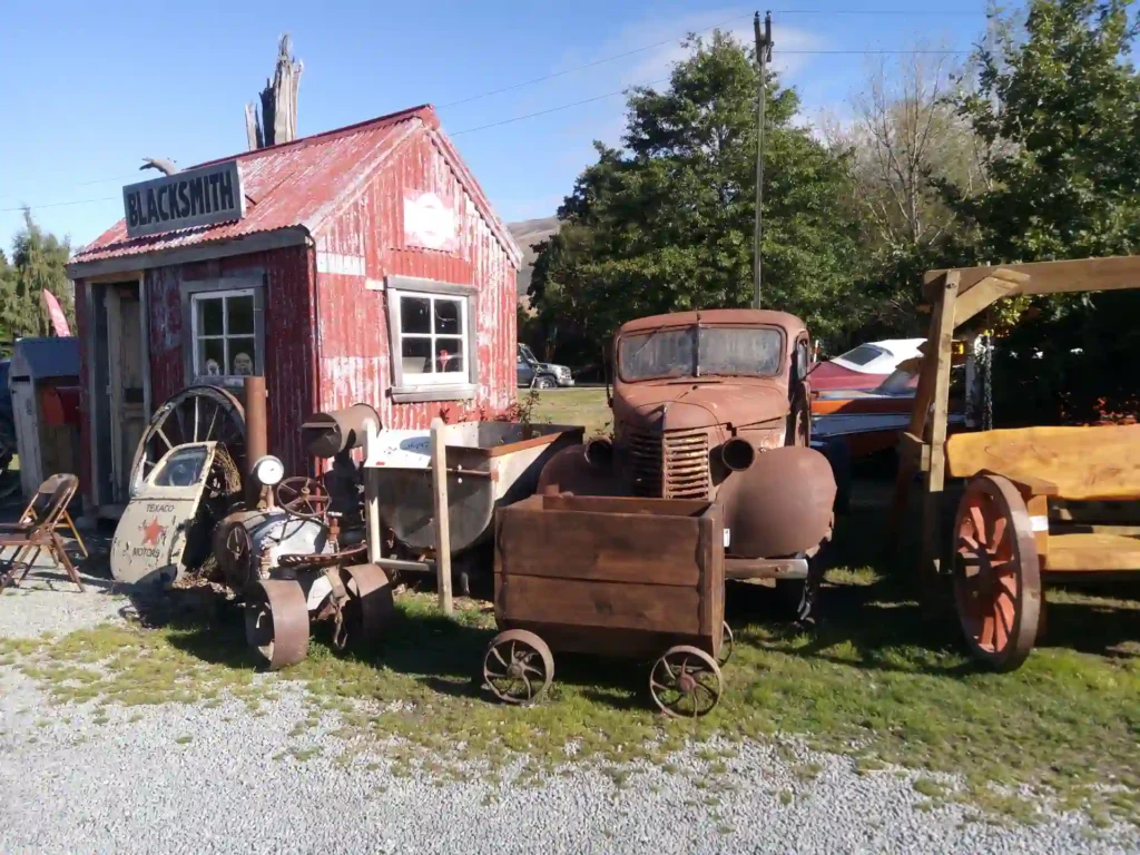 Eine rustikale Schmiede mit verblasstem roten Äußeren steht hinter alten, verrosteten landwirtschaftlichen Geräten, einem alten Lastwagen, Holzkarren und einem Wagenrad auf einer Wiese unter dem klaren blauen Himmel der Neuseeländischen Südinsel.