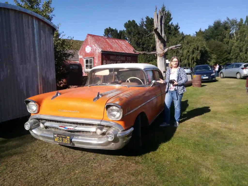 Eine Frau steht lächelnd neben einem orangefarbenen Chevrolet-Oldtimer, der auf einer Wiese auf der neuseeländischen Südinsel geparkt ist, mit anderen Autos und rustikalen Gebäuden im Hintergrund an einem sonnigen Tag.
