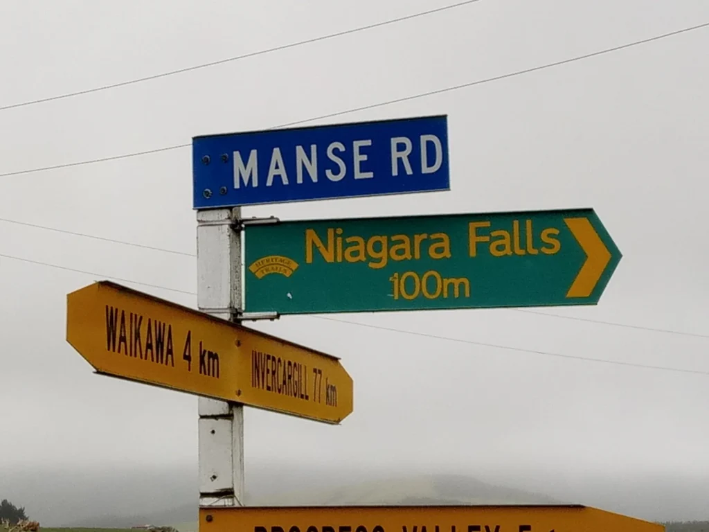 Straßenschilder an einer ländlichen Kreuzung auf der neuseeländischen Südinsel zeigen die Manse Rd und die Richtungen zu den Niagarafällen (100 m), Waikawa (4 km) und Wyndham (17 km) bei bewölktem Himmel an.