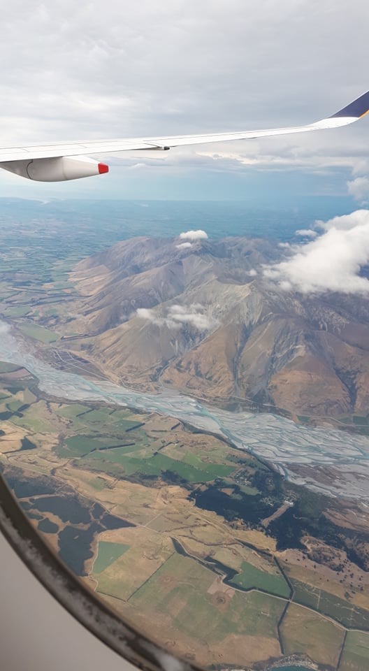 Luftaufnahme aus dem Fenster eines Flugzeugs, das eine Tragfläche über der Neuseeländischen Südinsel, gewundenen Flüssen und bunten Feldern unter einem bewölkten Himmel zeigt.