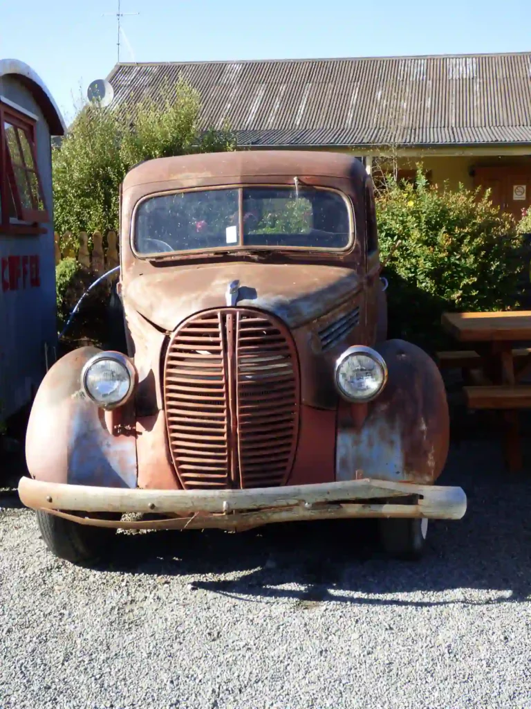 Ein rostiger Oldtimer-Lkw mit runden Scheinwerfern und einem großen Kühlergrill steht auf der landschaftlich reizvollen Neuseeländischen Südinsel auf einem Schotterplatz neben einer Holzbank und einem Schuppen, umgeben von Büschen und einem Gebäude.