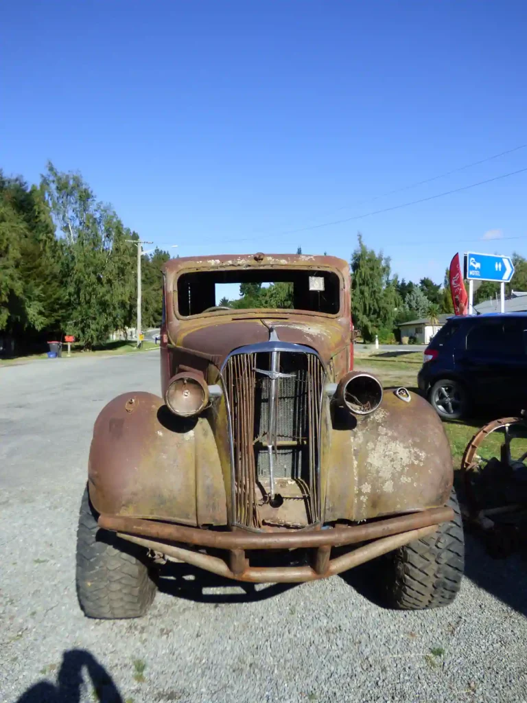 Ein rostiger, alter und teilweise demontierter Oldtimer mit fehlenden Scheinwerfern und Windschutzscheibe steht auf einem Schotterplatz unter blauem Himmel auf der Südinsel Neuseelands, mit Bäumen und anderen Fahrzeugen im Hintergrund.