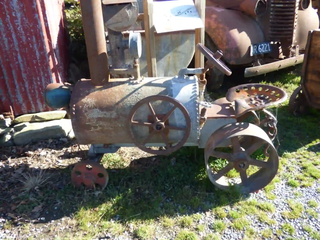Ein kleiner, verrosteter Oldtimer-Metalltraktor mit großen Speichenrädern und Traktorsitzen sitzt im Freien auf Gras und Kies in der Nähe von alten Fahrzeugen und einer Wellblechwand und erinnert an den ländlichen Charme der neuseeländischen Südinsel.