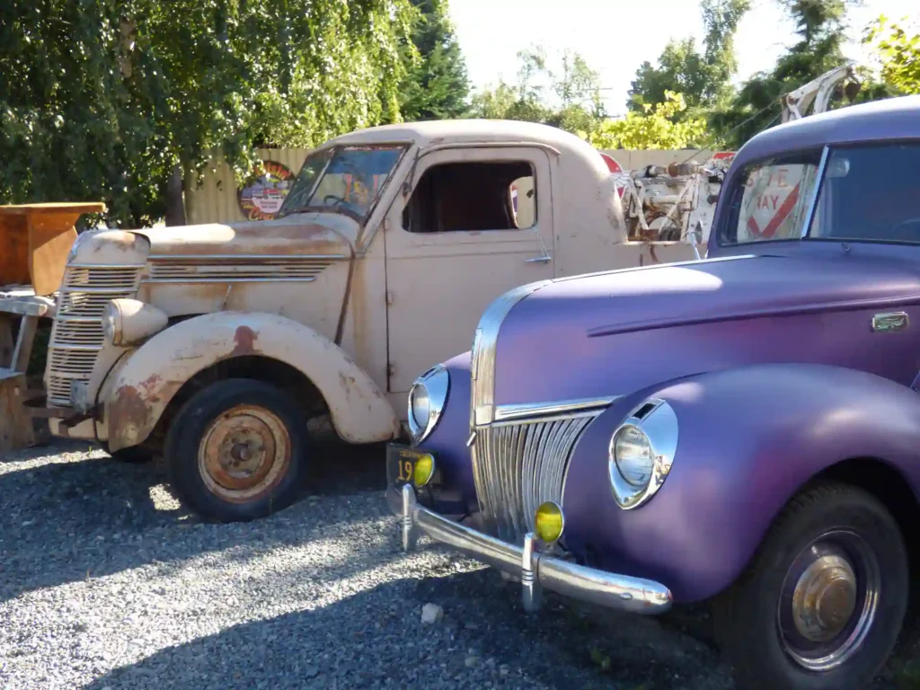 Zwei Oldtimer, die im Freien auf der neuseeländischen Südinsel geparkt sind: ein rostiger beiger Pickup und ein lila glänzender Oldtimer mit Chromdetails, beide auf einer Schotterfläche, umgeben von Bäumen und Sonnenlicht.