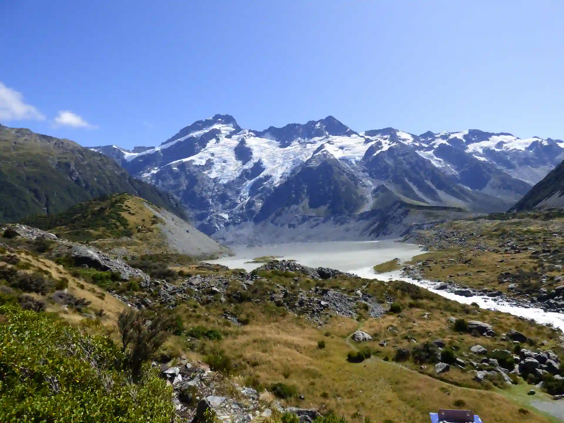 Schneebedeckte Berge unter blauem Himmel auf der neuseeländischen Südinsel, mit einem felsigen, grasbewachsenen Tal und einem Fluss im Vordergrund, der durch die Landschaft fließt.