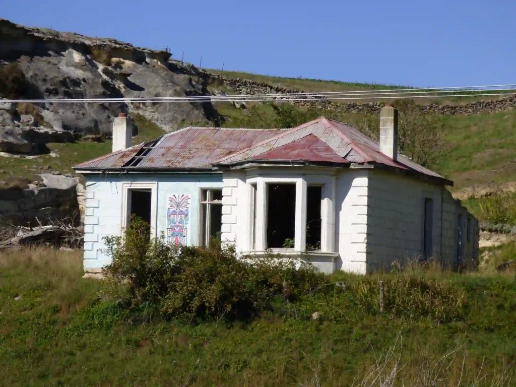 Ein altes, verlassenes weißes Haus mit verrostetem rotem Dach und zerbrochenen Fenstern steht auf einer überwucherten Wiese auf der neuseeländischen Südinsel, im Hintergrund sieht man Hügel und Felsen unter einem klaren blauen Himmel.