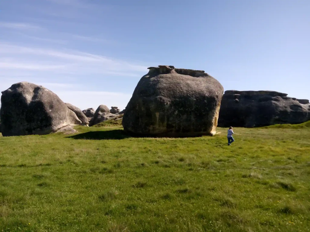 Eine Person geht über eine grasbewachsene Wiese auf der neuseeländischen Südinsel, mit großen, abgerundeten Felsbrocken im Hintergrund unter einem klaren blauen Himmel.