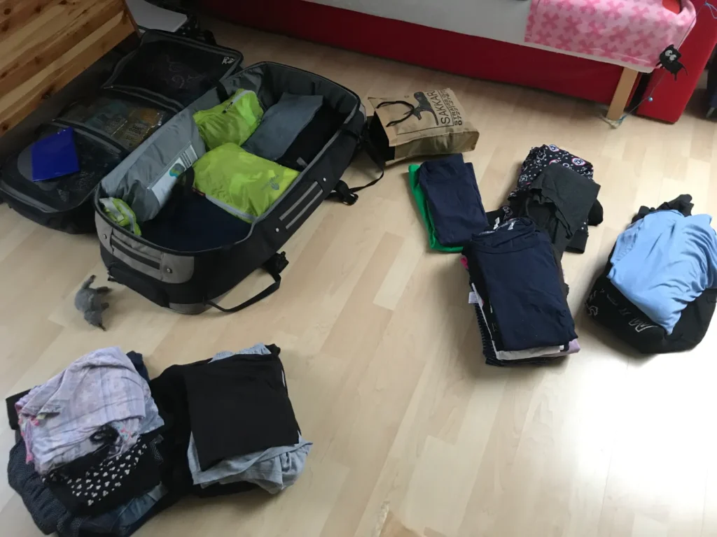 Ein halb gepackter Koffer auf Holzboden, daneben ordentliche Kleiderstapel und eine Tüte; dahinter ein Bett mit rosa Decke.