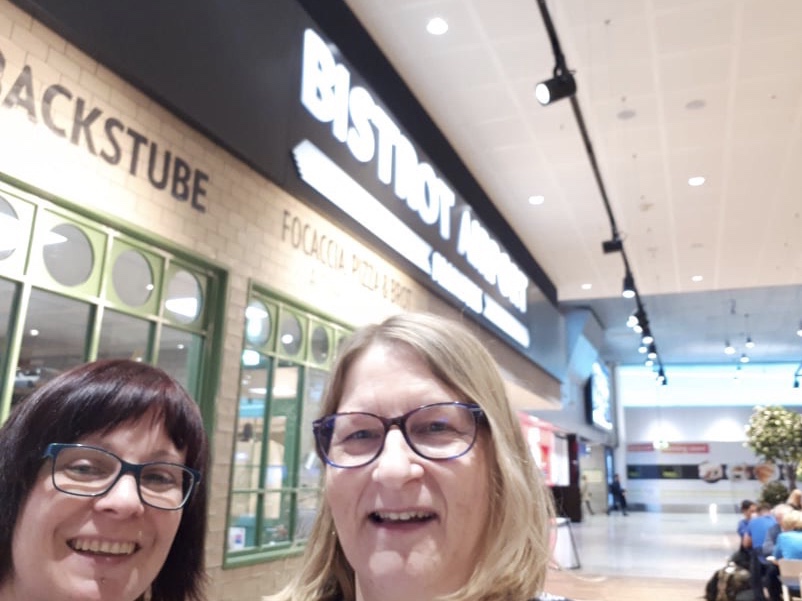 Zwei Frauen mit Brillen lächeln für ein Selfie in einem hellen Flughafenterminal neben einem Schild mit der Aufschrift BISTROT AIRPORT. Im Hintergrund sind grün umrahmte Fenster und andere Reisende zu sehen, die auf dem Weg zur Neuseeländischen Südinsel sind und auf bevorstehende Abenteuer hinweisen.